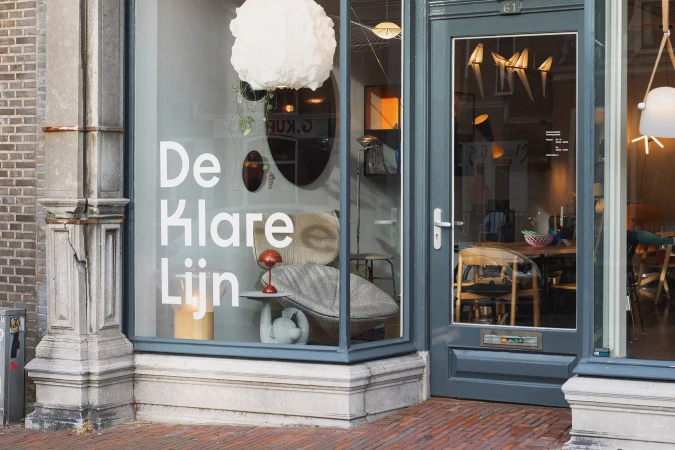 Storefront window of a shop displaying furniture, lighting, and decor with  'De Klare Lijn' text on the window. Gray exterior and brick sidewalk.