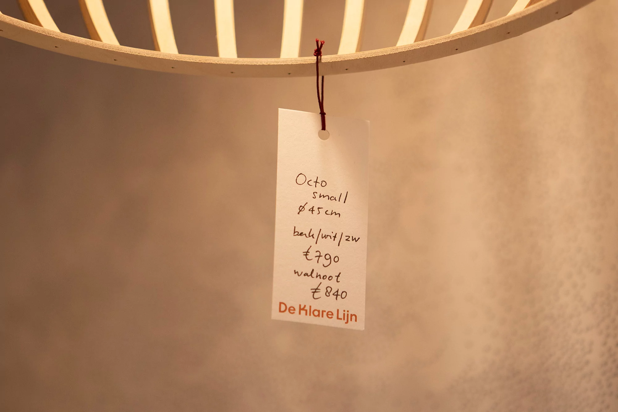 Close-up of a hanging light fixture with a tag displaying pricing details. The light's wooden structure is visible against a neutral background.