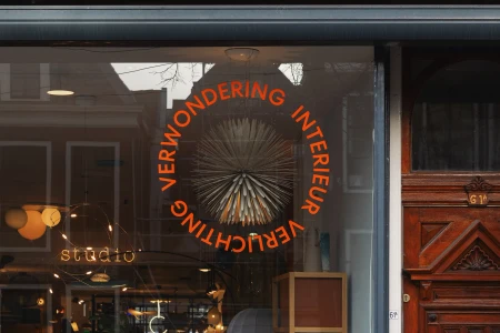 Storefront window of De Klare Lijn displaying bold circular orange typography and an interior lighting fixture.