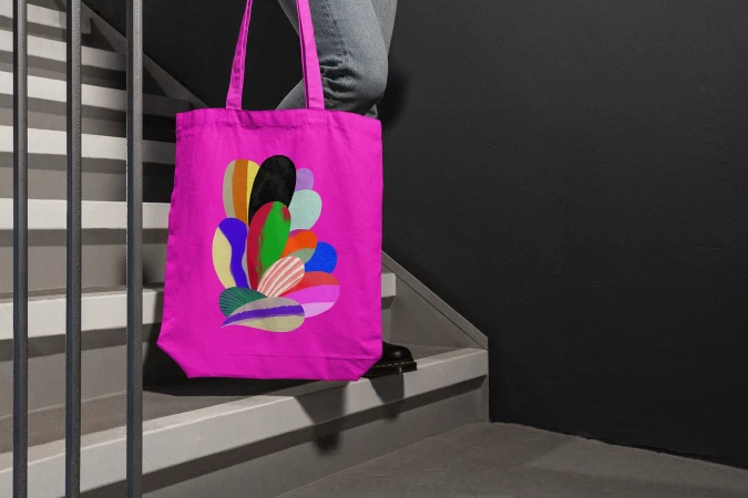 Bright pink tote bag with colorful abstract design on person standing on grey stairs against a dark wall, showcasing fashion accessory.