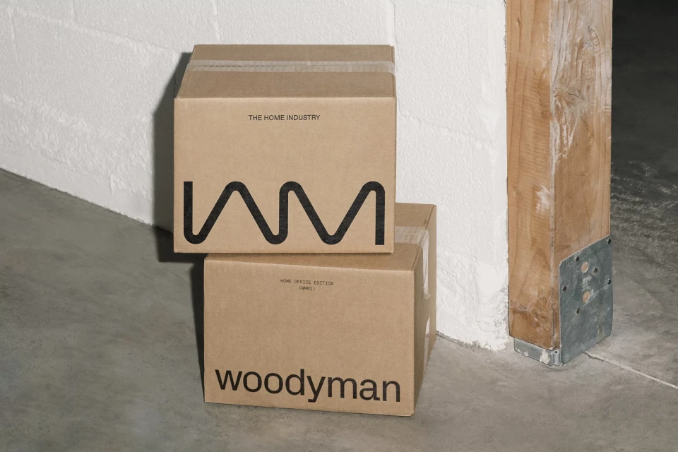 Two stacked cardboard boxes labeled 'The Home Industry' and 'Woodyman' sit indoors against a white brick wall and a wooden post.