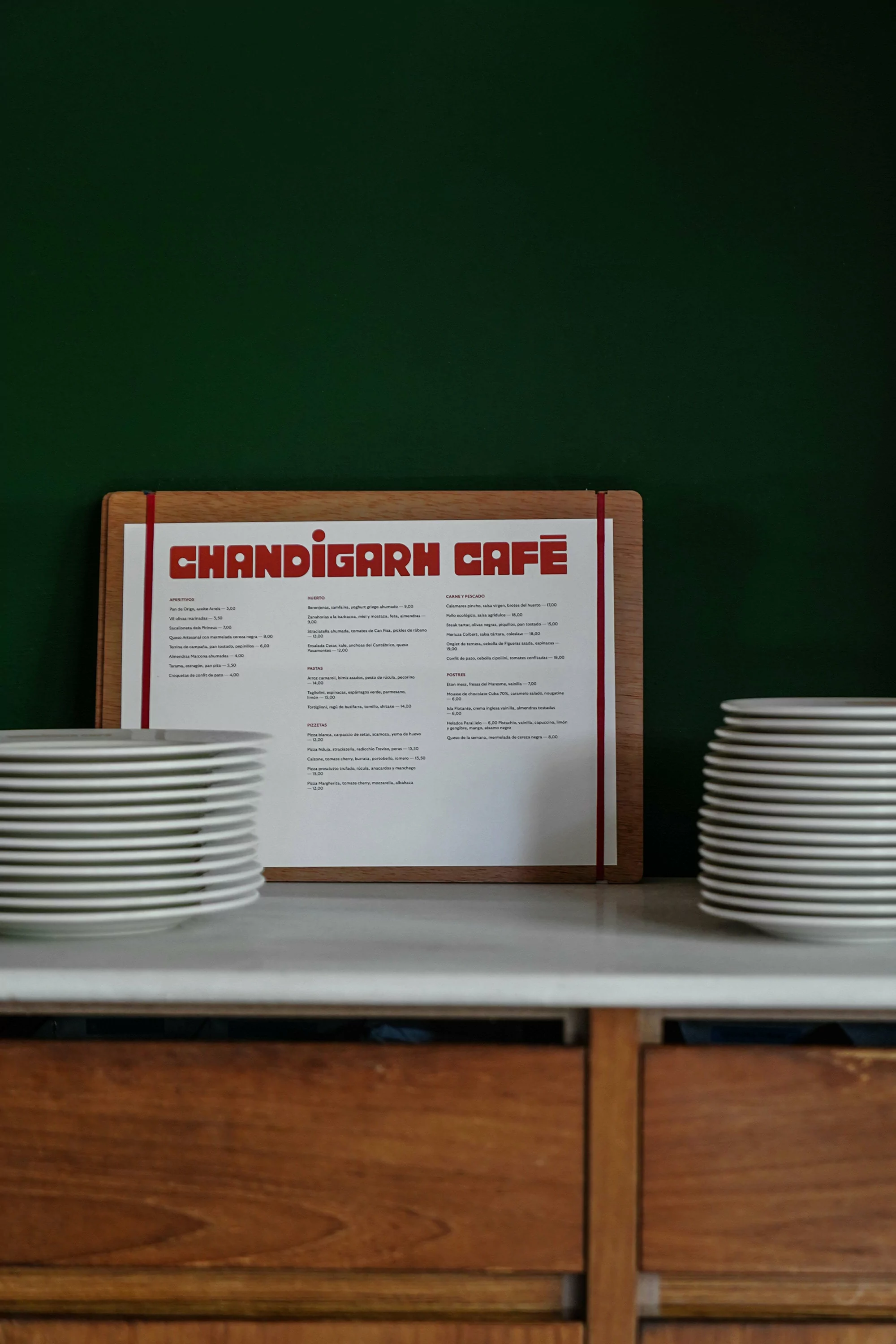 Cafe menu board on a white counter between two stacks of plates against a dark green wall and wood cabinetry.