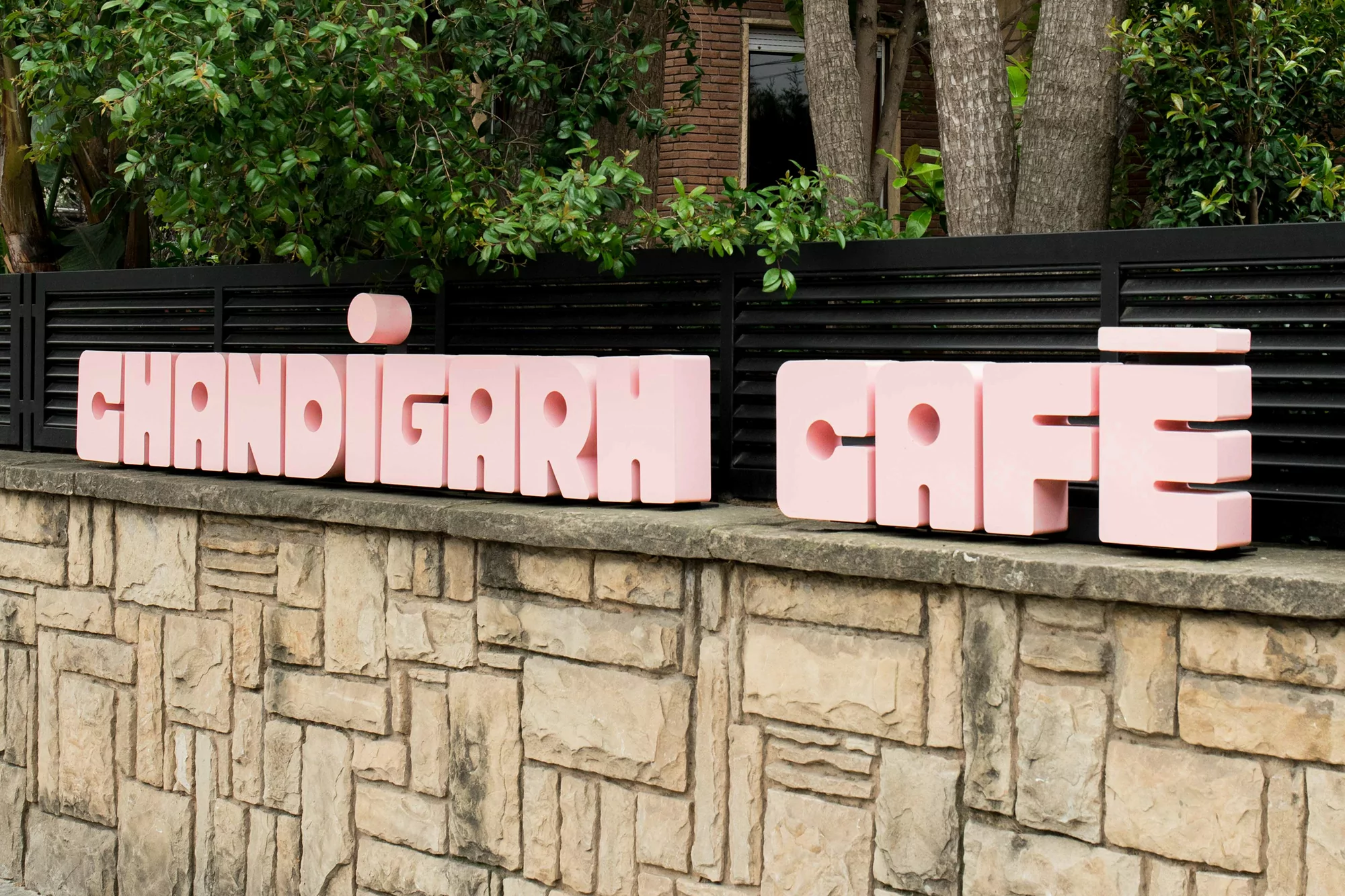 Pink Chandigarh Cafe signage on a stone wall, with green foliage and a black fence in the background, creating a modern outdoor cafe display.