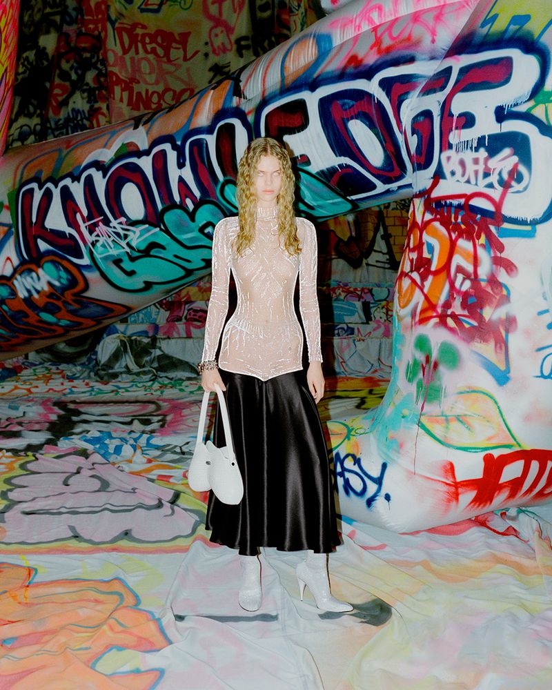 Woman in a light top and black skirt poses in front of graffiti art. She holds a white bag and wears matching white heeled boots.