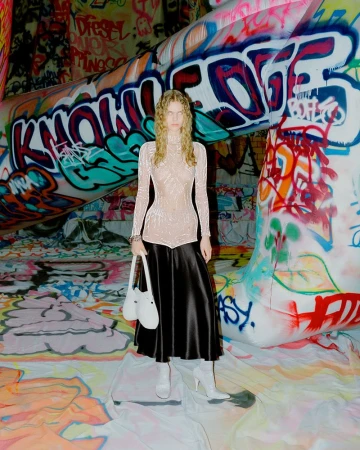 Woman in a light top and black skirt poses in front of graffiti art. She holds a white bag and wears matching white heeled boots.