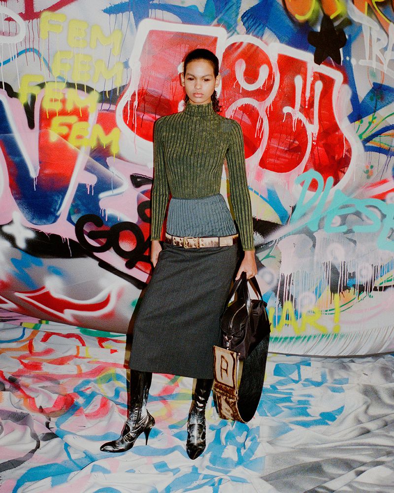 A woman poses in a studio wearing a dark skirt, green sweater, and high-heeled boots, holding a handbag, with a graffiti background.