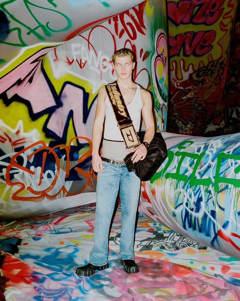 Male fashion model poses indoors against a graffiti-covered backdrop, wearing a white tank top, jeans, and black platform shoes.
