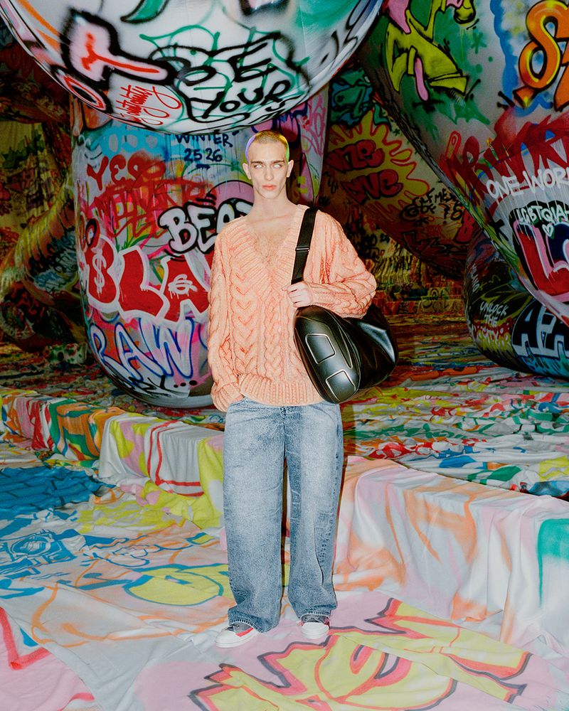 Young adult stands in a peach sweater, light-wash jeans, and black bag, against a backdrop of graffiti-covered structures and floor.