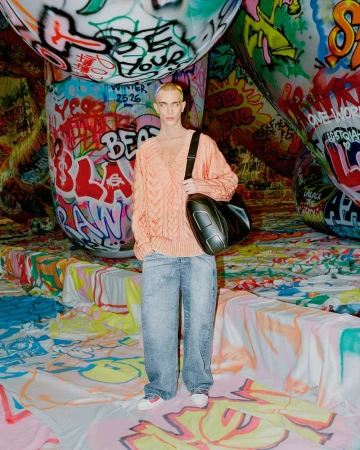 Young adult stands in a peach sweater, light-wash jeans, and black bag, against a backdrop of graffiti-covered structures and floor.
