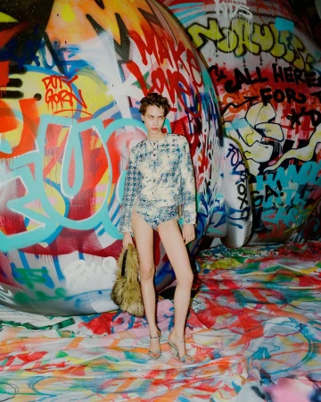 A fair-skinned woman in patterned top and bottoms poses in front of graffiti-covered wall with a colorful floor covering and furry handbag.