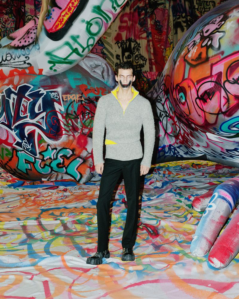 Full-length portrait of a man in a sweater and black pants standing indoors surrounded by graffiti and colorful objects and patterns.