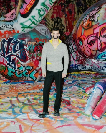 Full-length portrait of a man in a sweater and black pants standing indoors surrounded by graffiti and colorful objects and patterns.