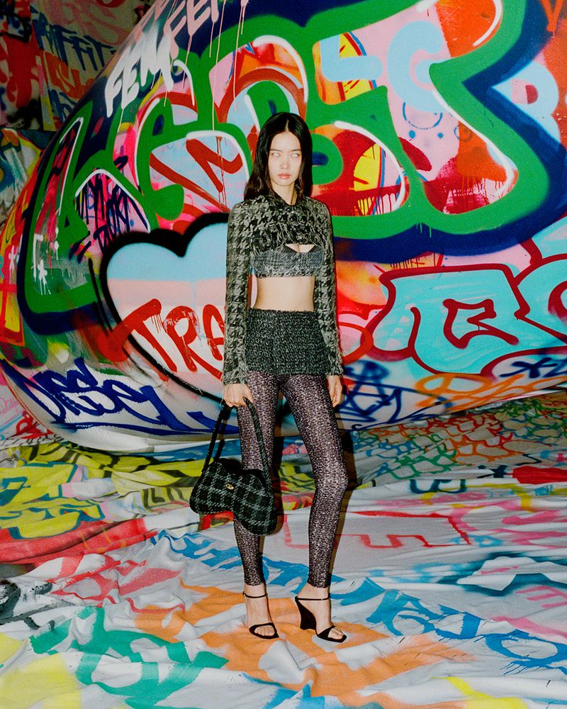 Fashion model poses against graffiti-covered surface in a cropped houndstooth jacket, black mini skirt, patterned leggings, and black heels.