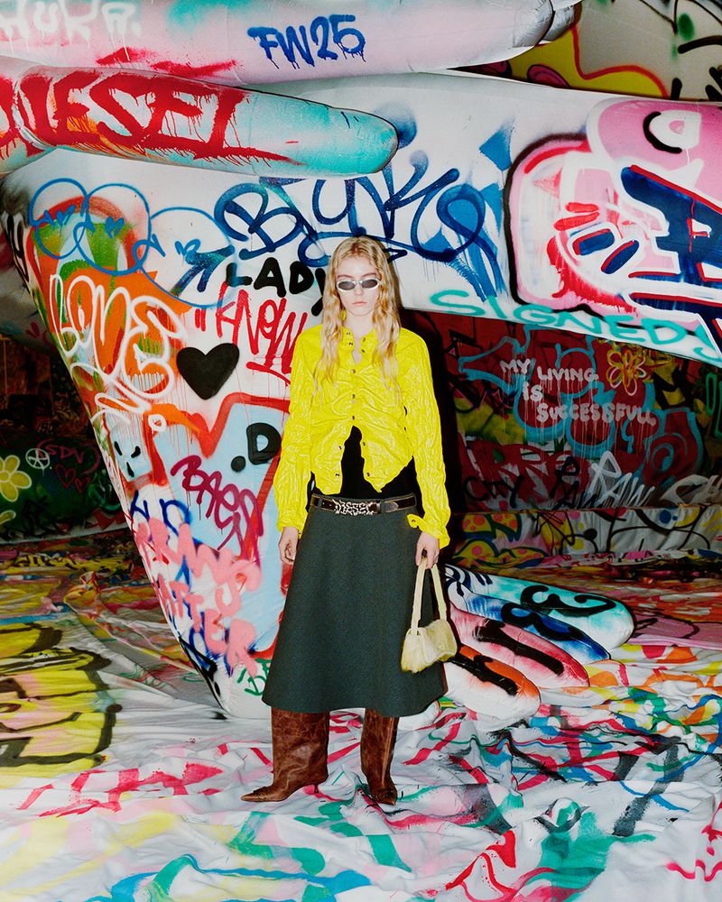 A woman poses in a yellow cardigan, dark green skirt, and brown boots in front of a graffiti-covered backdrop, holding a small purse.