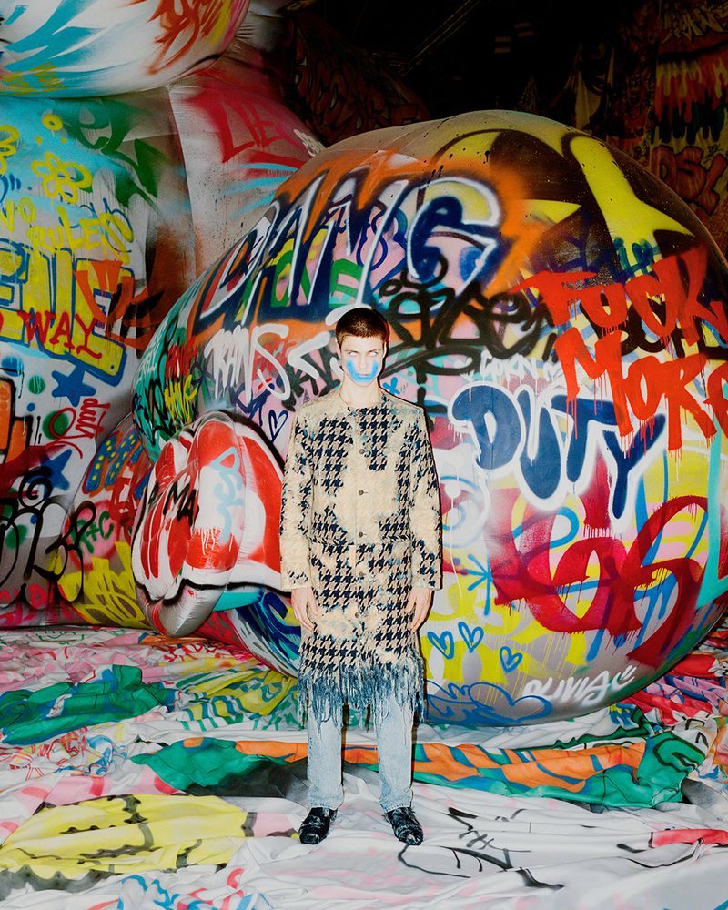Adult male model poses in a fringed houndstooth coat and light denim jeans against a graffiti-covered backdrop in an indoor setting.