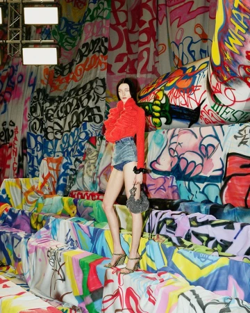 A slender woman posing in a red jacket and denim shorts, stands amidst a vibrant, graffiti-style indoor setting.