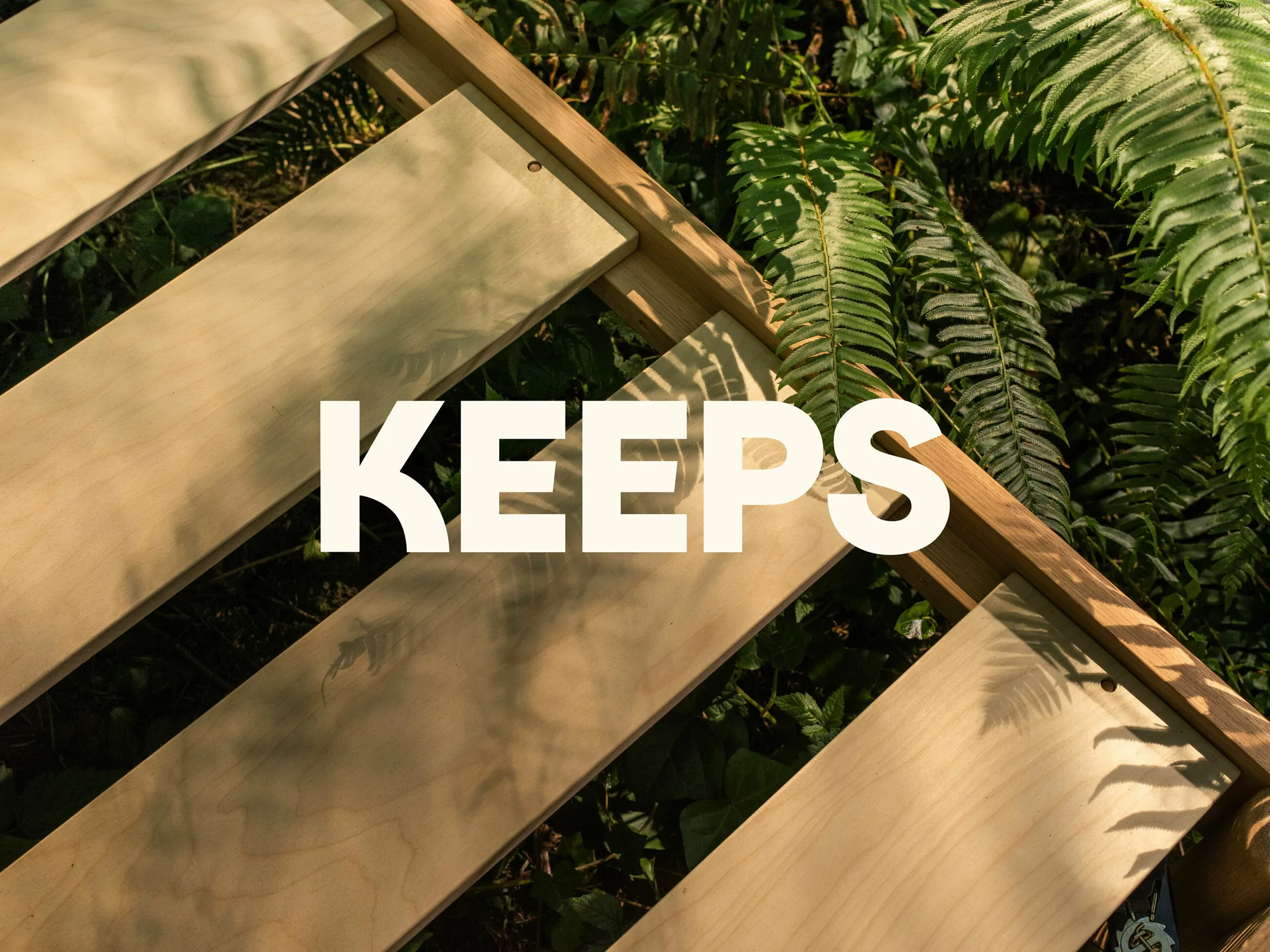 Wooden bed frame is angled on a dark green fern background with overlaid white text that reads “KEEPS” in bold font.