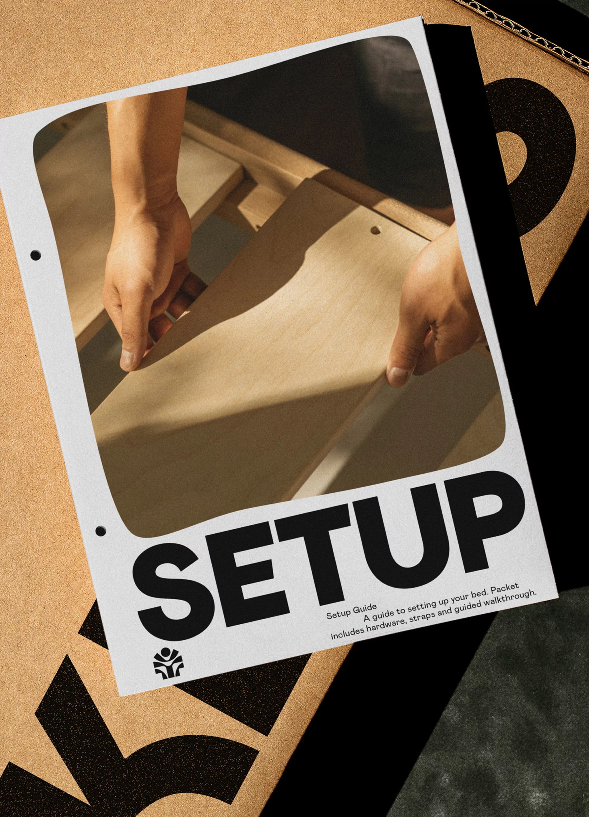 Assembly guide lying on cardboard surface. Hands appear in frame, positioning plywood panel. Set-up instructions detail hardware, straps.