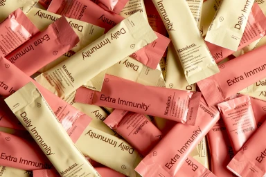 Manukora supplement stick packs in pale cream and pink scattered flat lay against a matching background.