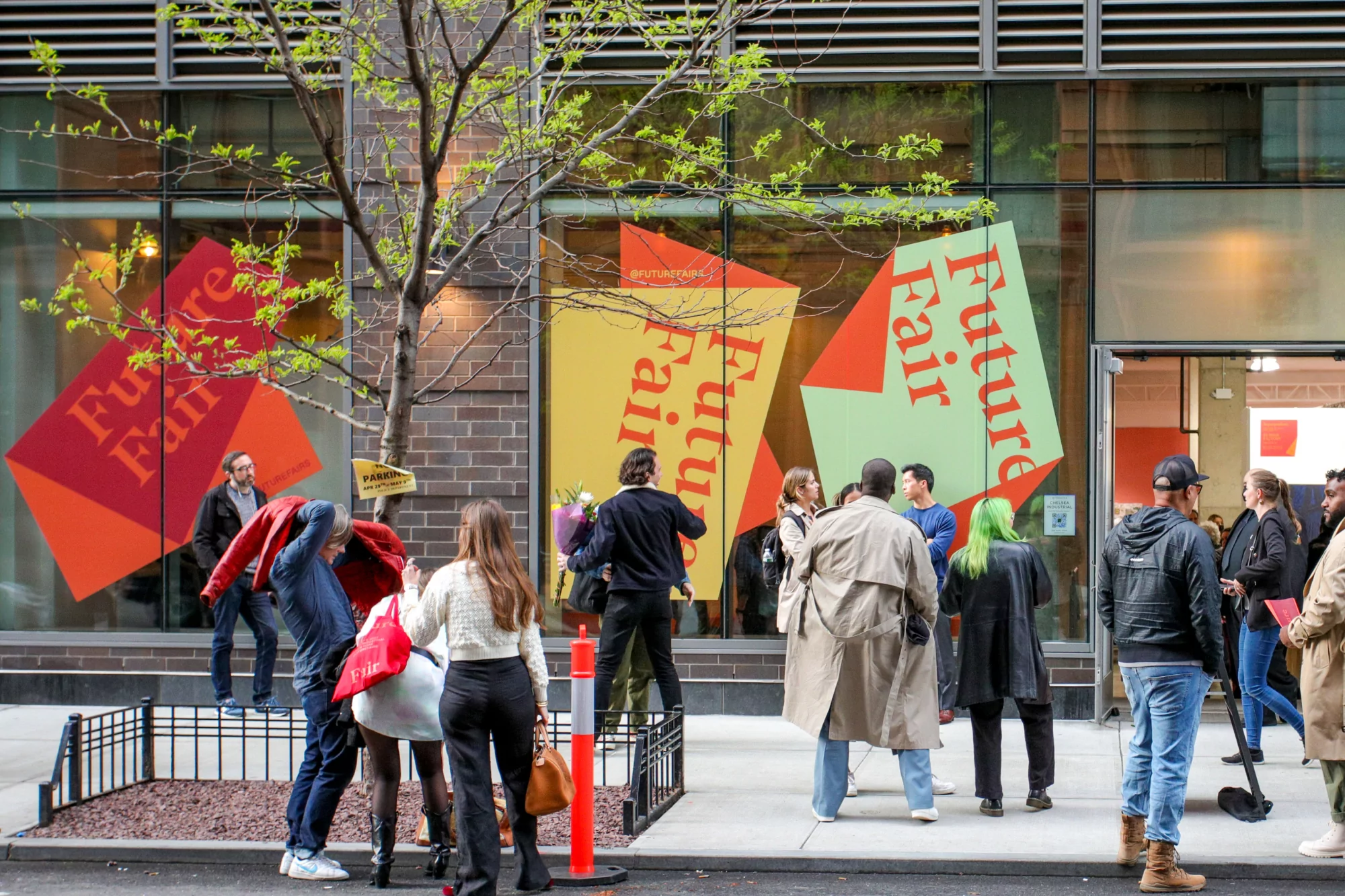 People gather near the entrance of an event venue with large signage, under a tree with budding leaves, on an overcast day in an urban setting.