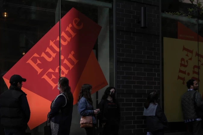 People stand near a building in front of posters for Future Fair, with text visible; an outdoor scene with architectural details.