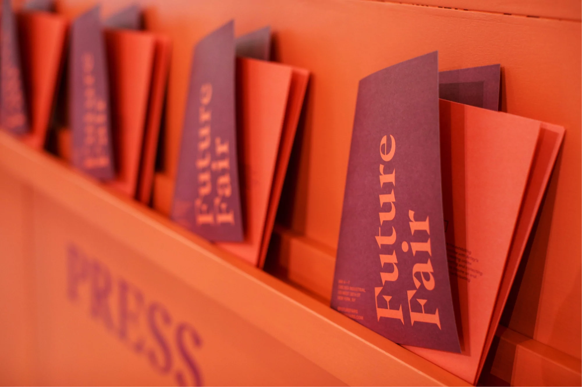 Future Fair brochures displayed on an orange rack, showcasing event information in a close-up, studio setting with muted lighting.