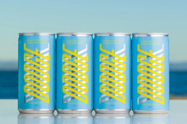 Four light blue beverage cans featuring yellow typography arranged in a row against an outdoor horizon backdrop.