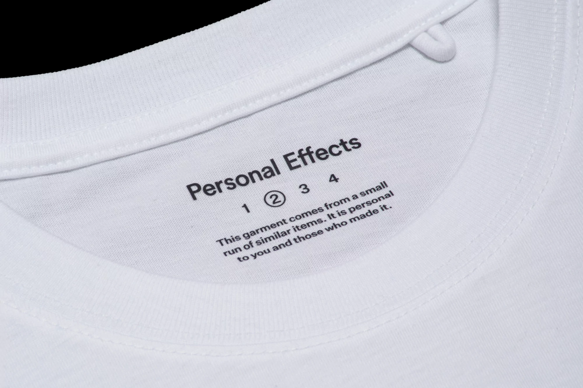 Close-up of a white t-shirt showing the label and collar.  The label reads 'Personal Effects' and text indicating a small run of items.