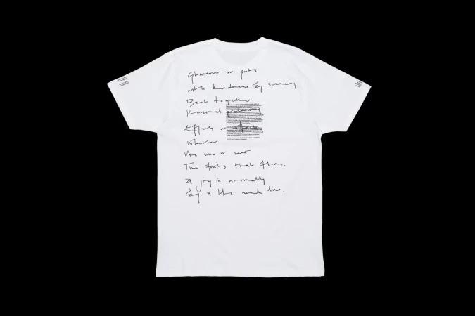 A white t-shirt on a black background featuring dark calligraphic-style text arranged asymmetrically over the garment's surface.