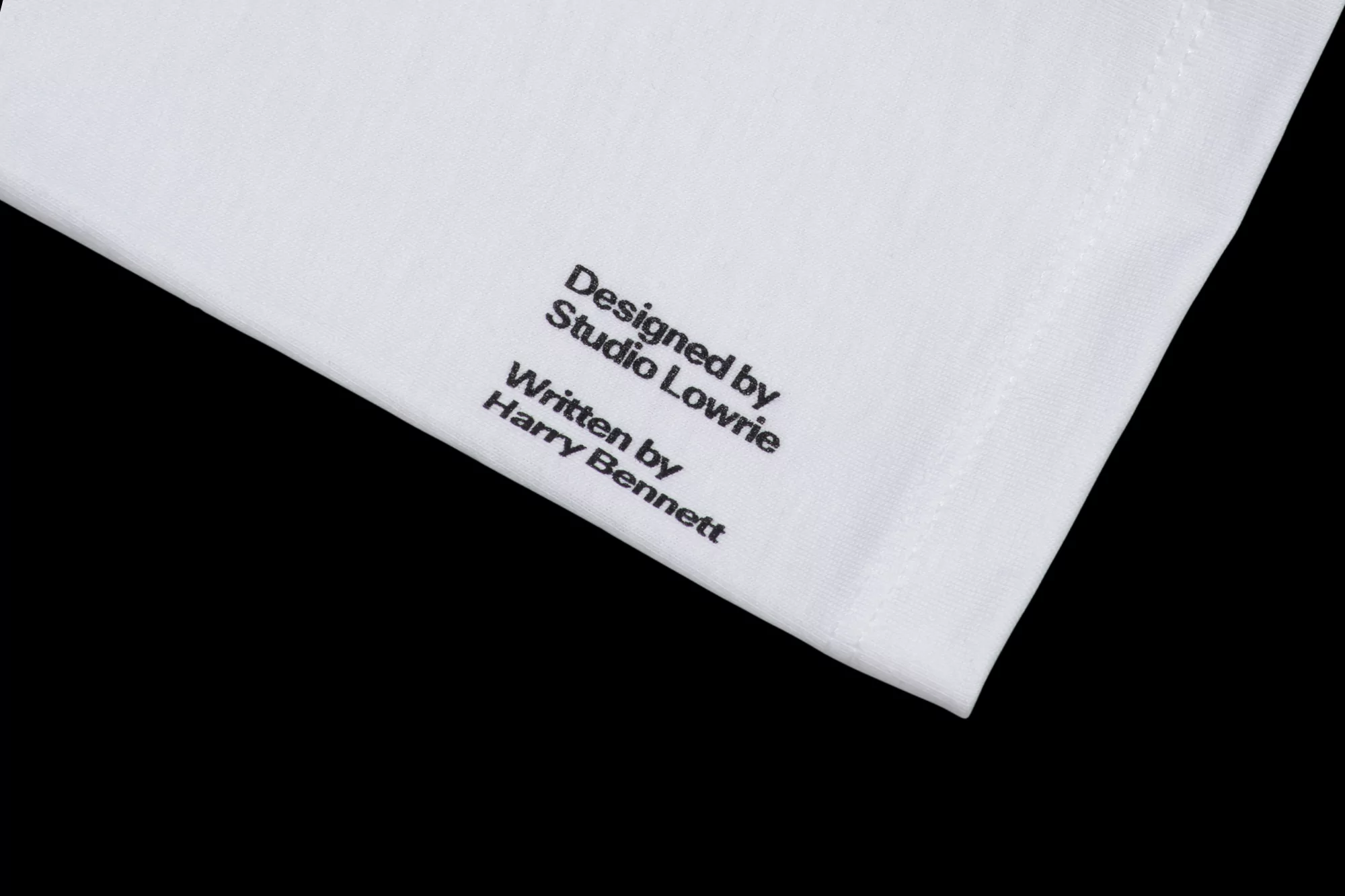 Close-up of a white cloth with black text that reads 'Designed by Studio Lowrie, Written by Harry Bennett' against a black background.