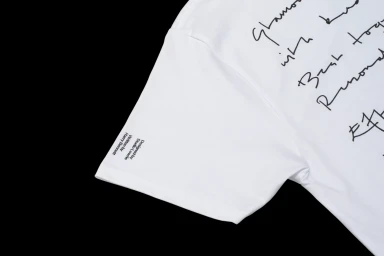 Close-up of a white t-shirt featuring black handwritten script and small credited typography on the sleeve.