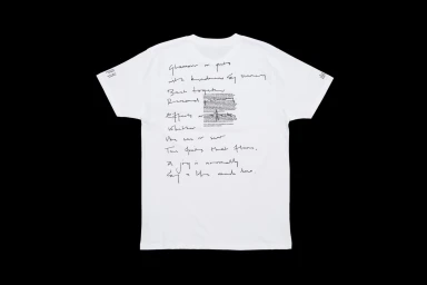 White t-shirt displayed flat against a black background featuring handwritten cursive poetry and a printed text block on the back