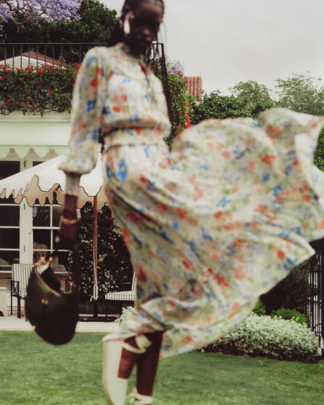 Woman in a floral dress with a handbag poses in front of a white building surrounded by green grass and trees on a cloudy day.