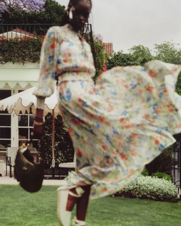 Woman in a floral dress with a handbag poses in front of a white building surrounded by green grass and trees on a cloudy day.