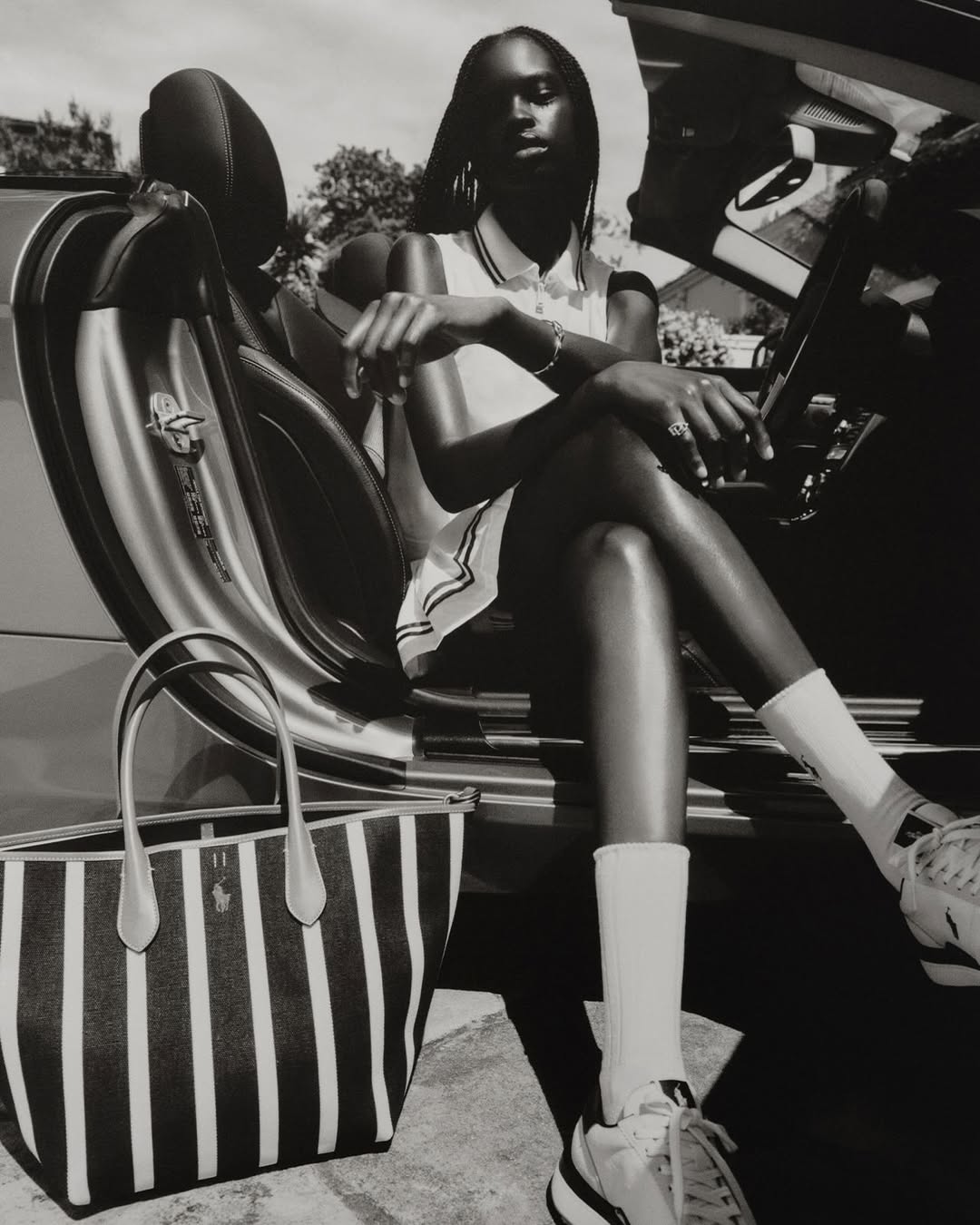 Black and white portrait of a dark-skinned woman sitting in a car with a striped tote bag, sneakers, and white socks in a monochrome style.