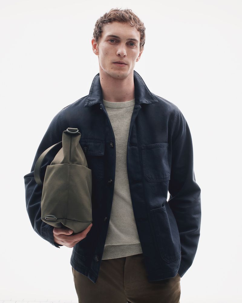 A man stands in a bright photo studio, wearing a navy blue jacket and brown pants, while holding a green tote bag in a fashion editorial shot.