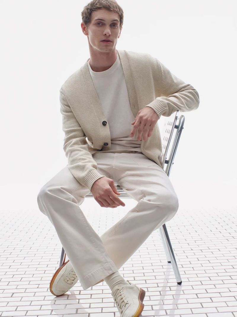A young adult man in a neutral-colored cardigan, t-shirt, pants, and sneakers sits on a chair with a white brick background.