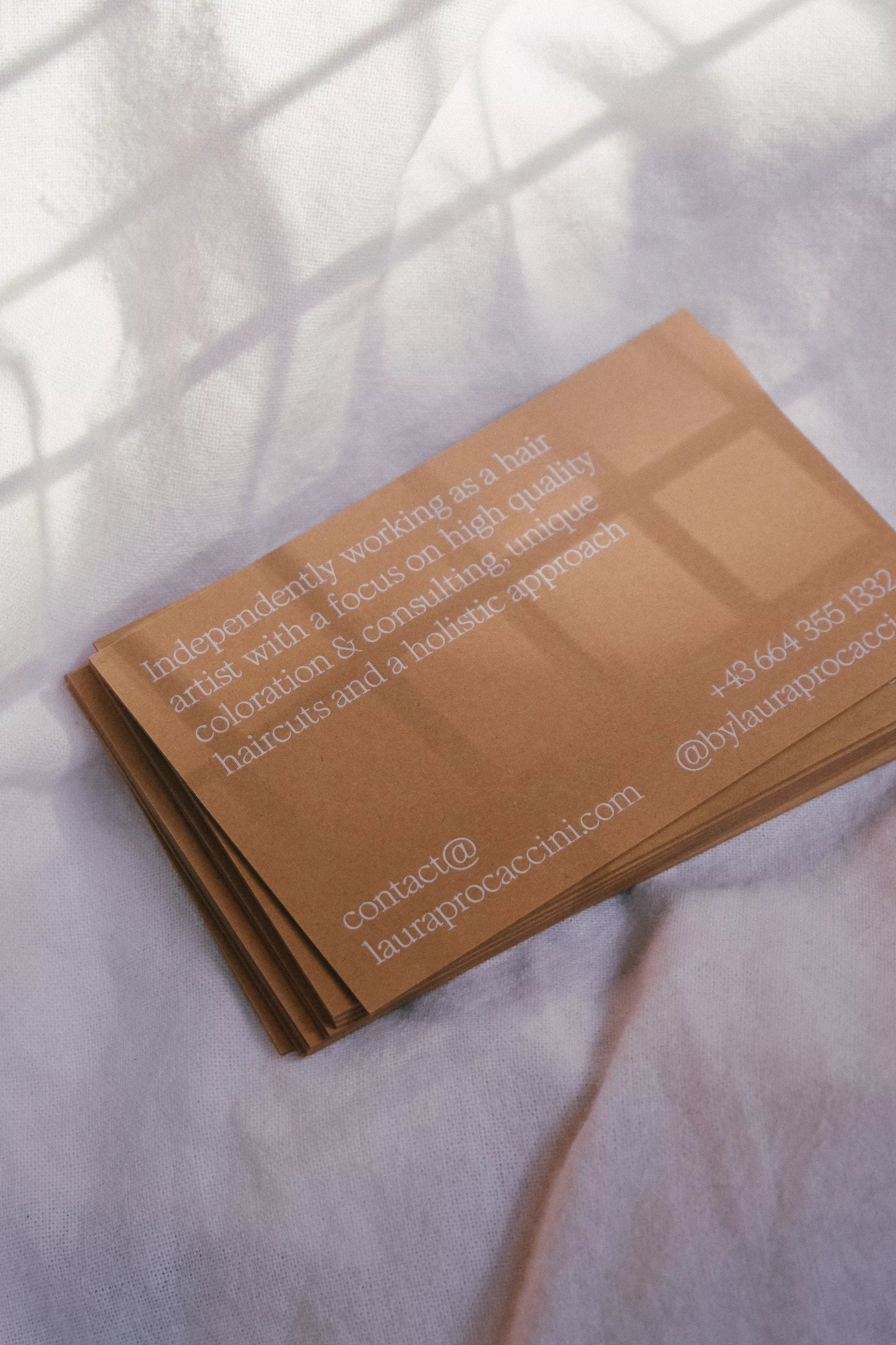 Tan-colored business cards with white text rest on a white fabric surface, light from a window creates a soft shadow pattern.