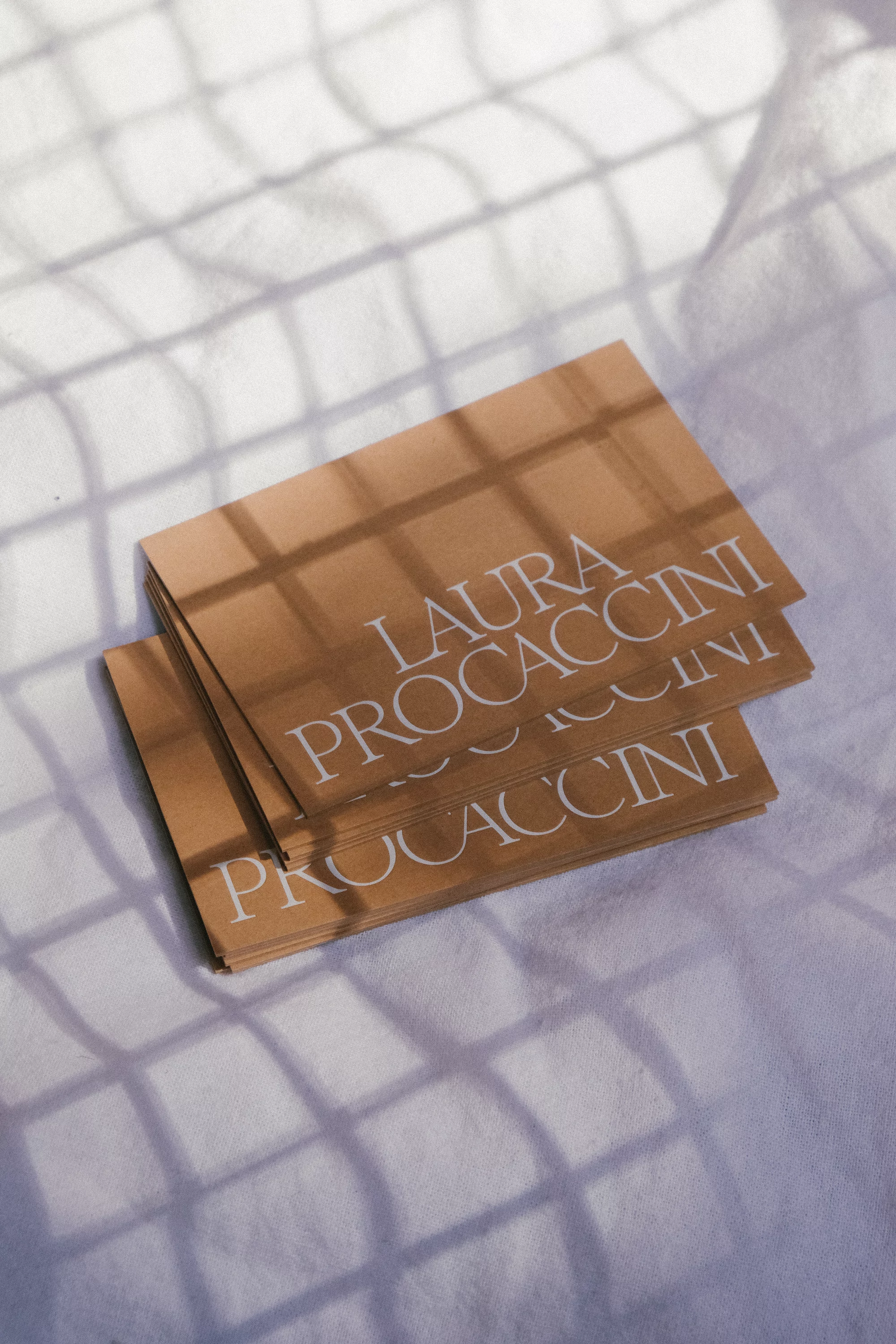 Stack of tan business cards with white text on a white textured surface with shadows, creating an elegant, minimalist aesthetic.