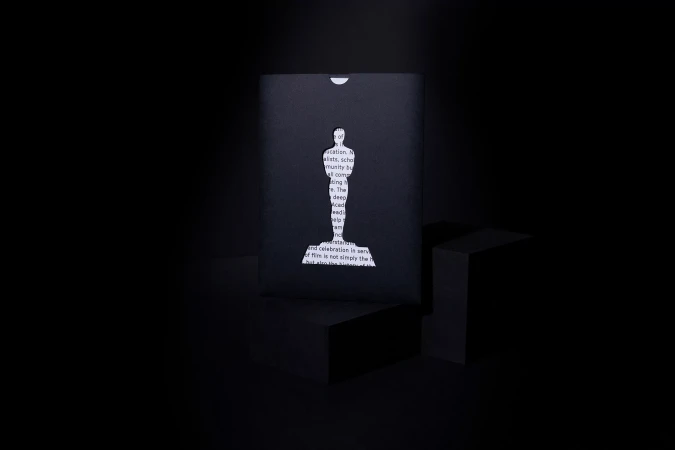 Minimalist presentation of a black envelope featuring a die-cut silhouette of a statuette, illuminated against a dark background.