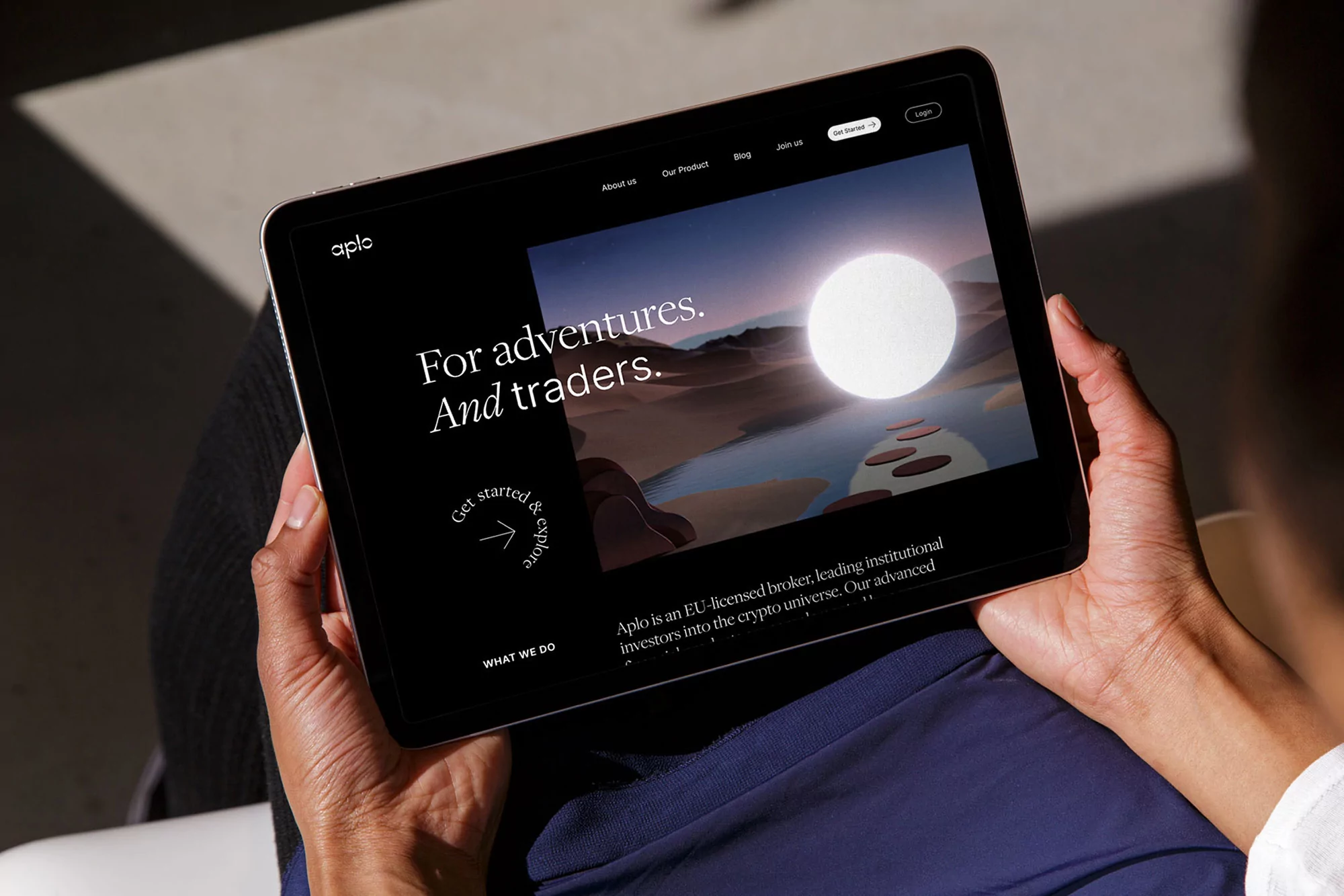 Person holding a tablet displaying the 'aplo' website with desert landscape and financial text, indoors with natural lighting and a modern design.