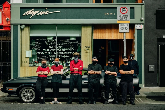 Group of masked restaurant employees posing in front of their establishment, with a vintage car parked beside the entrance.