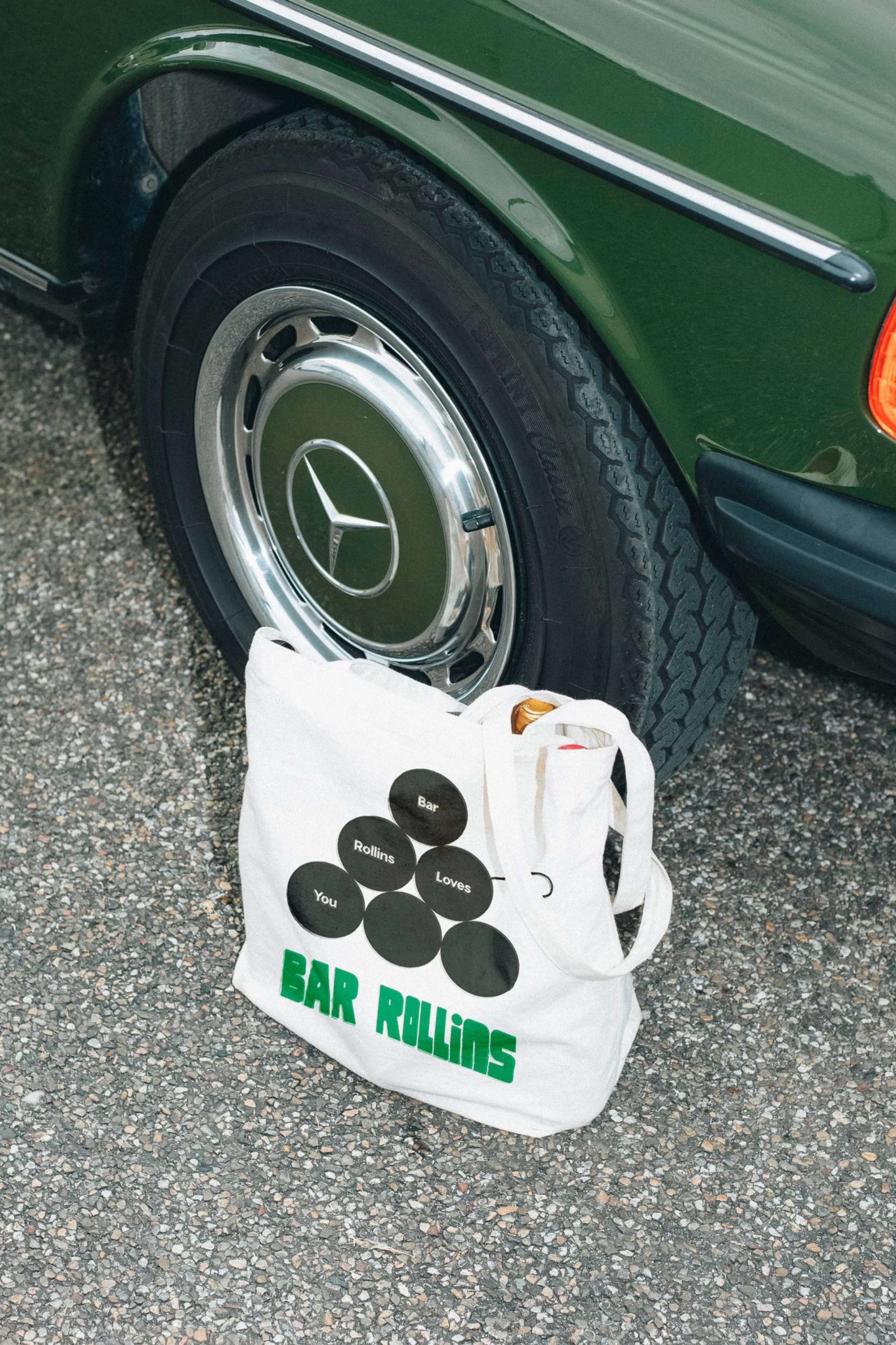 Tote bag with 'Bar Rollins' text next to vintage Mercedes car wheel, set against asphalt backdrop in a street setting.