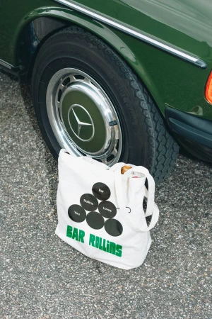 Tote bag with 'Bar Rollins' text next to vintage Mercedes car wheel, set against asphalt backdrop in a street setting.