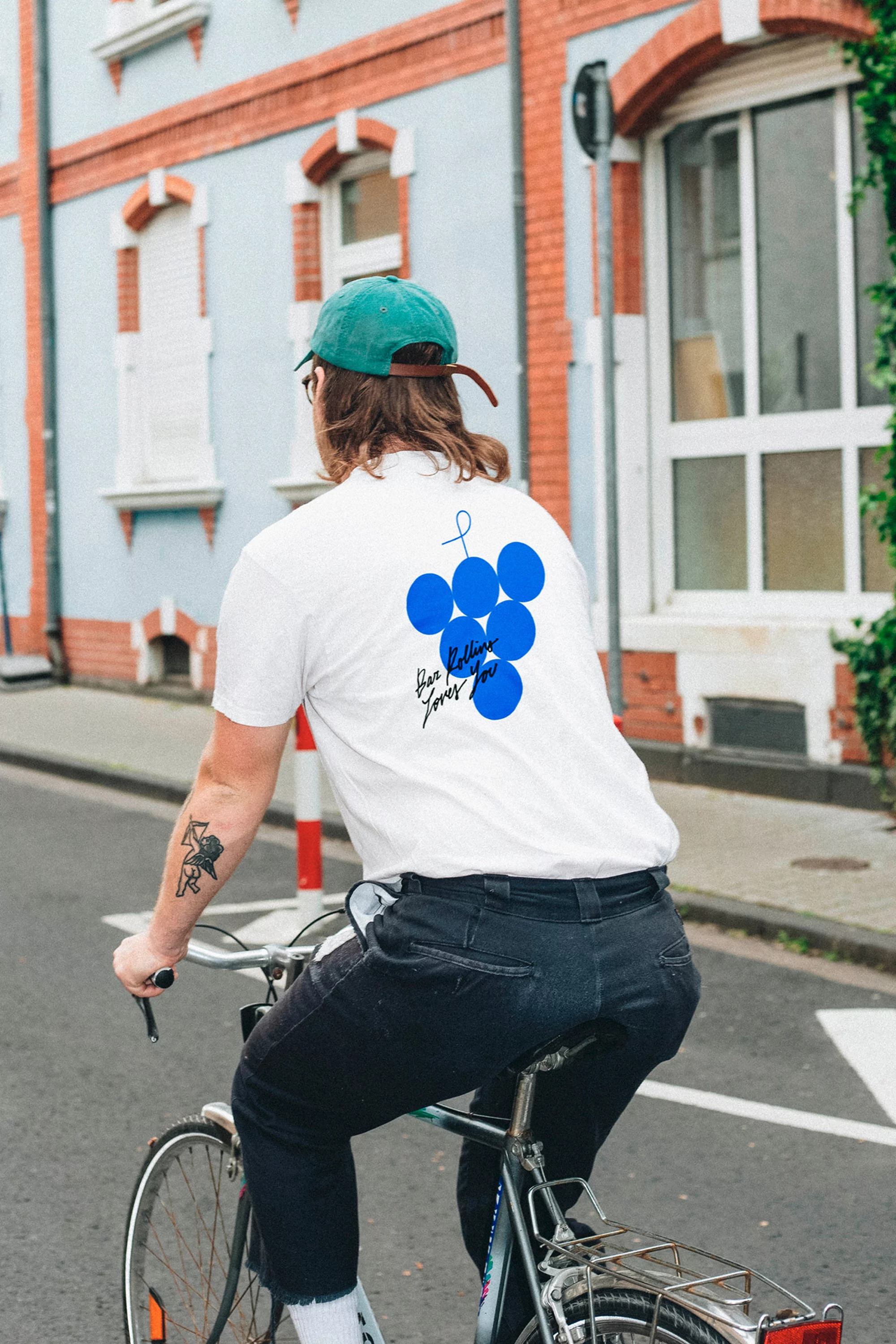 Back view of a man riding a bicycle on a city street, wearing a white t-shirt with a graphic design and a green baseball cap.