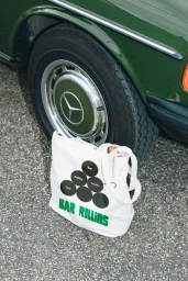 White canvas tote bag with Bar Rollins branding resting on asphalt in front of a green car tyre.