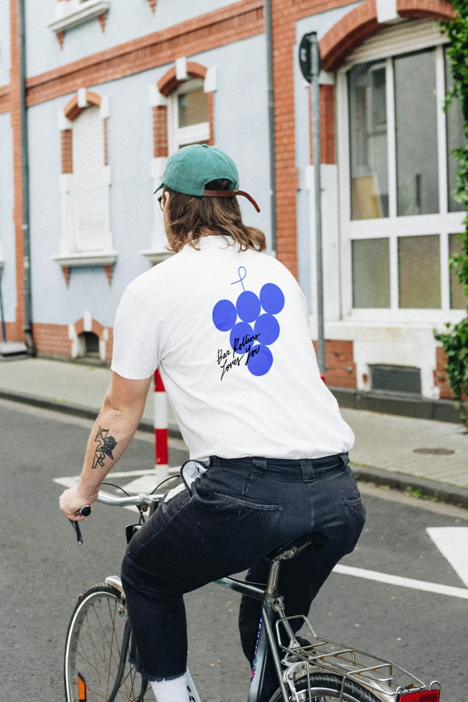Man riding a bicycle on a city street wearing a white t-shirt with a blue graphic, dark pants, and a green cap
