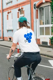 Man riding a bicycle on a city street wearing a white t-shirt with a blue graphic, dark pants, and a green cap
