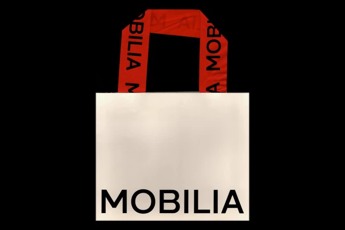 Close-up studio shot of a white shopping bag with a red handle printed with the word 'MOBILIA' against a black background.