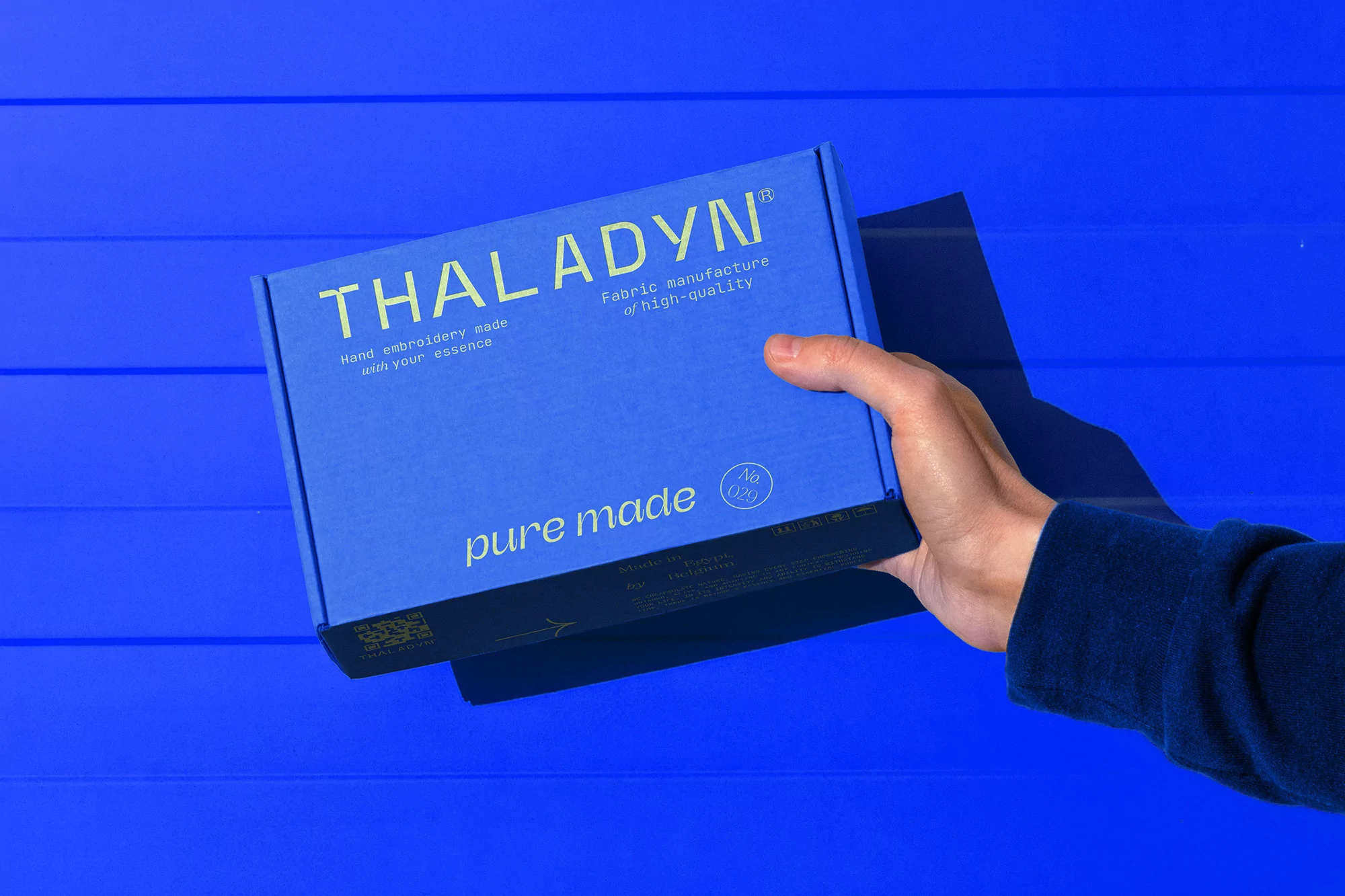 Hand holds a blue THALADYN fabric manufacture box against a blue wooden surface in a top-down shot with product details.